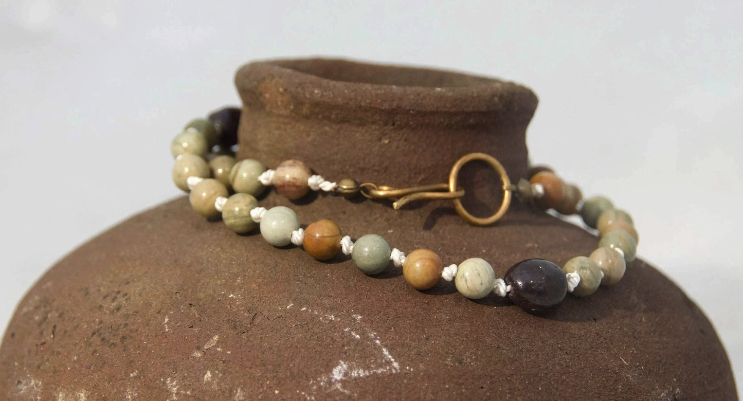 A beaded necklace with natural stone beads and a gold clasp draped over a rustic brown ceramic vase against a white background.