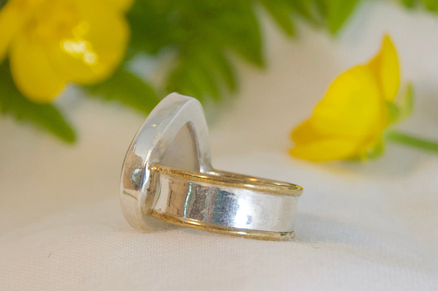 Silver ring on a white surface with yellow flowers and green fern leaves.