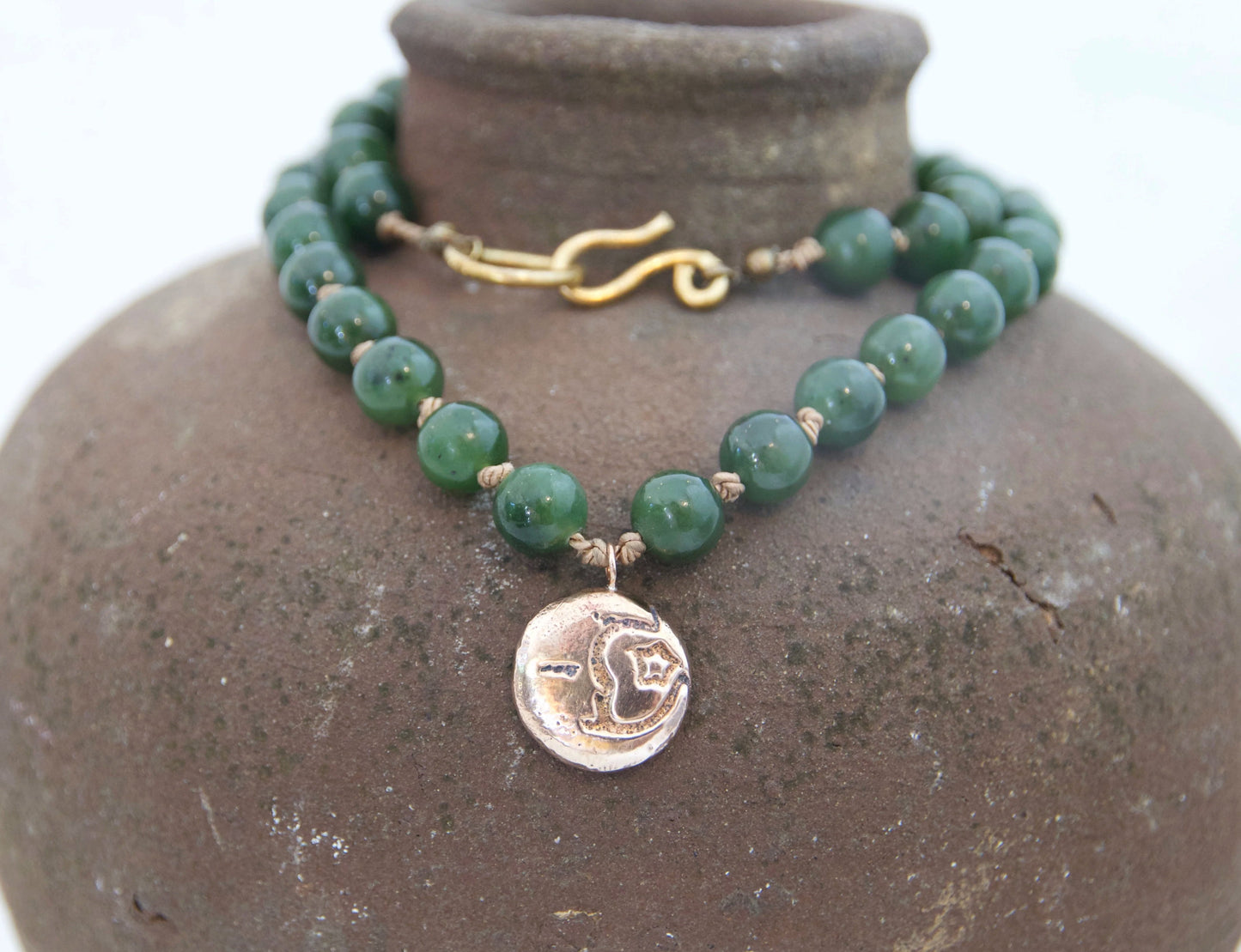 A beaded necklace with green round beads, a gold-tone toggle clasp, and a bronze circular pendant, displayed on a brown ceramic vase against a white background.