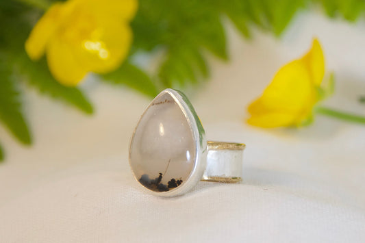 Silver ring with a teardrop-shaped stone on a white surface with green fern leaves and yellow flowers.