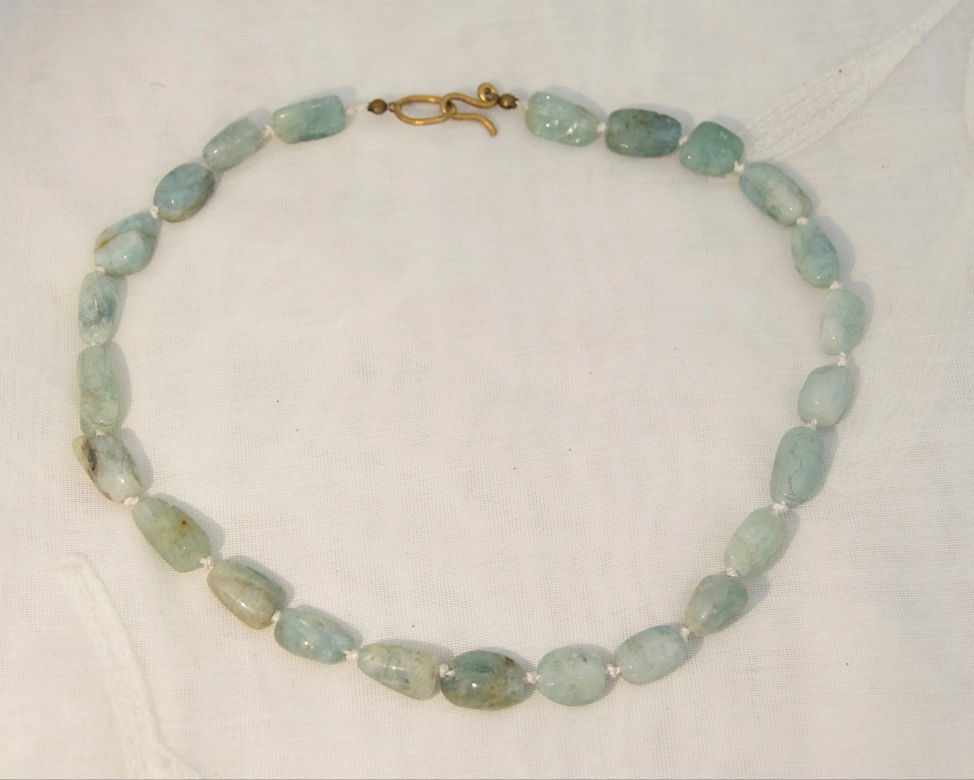 A beaded necklace made of light green oval beads with a small gold clasp, displayed on a white textured fabric background.