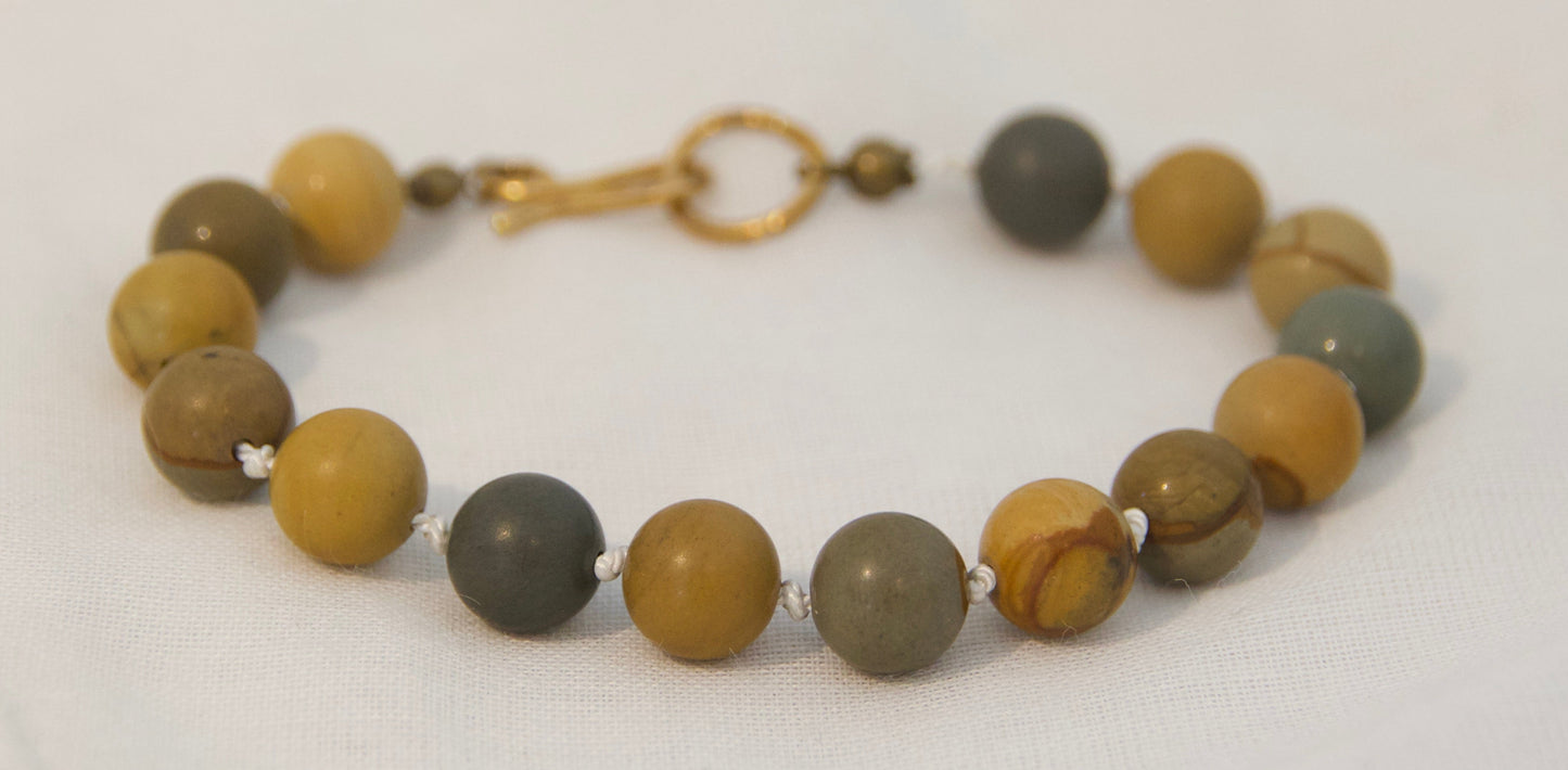 A beaded bracelet made of round stone beads in earthy brown, olive green, and mustard yellow tones, with a small gold clasp, resting on a white fabric background.