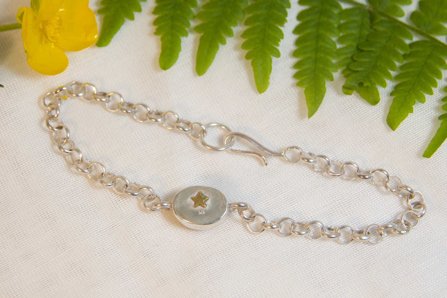 Silver bracelet with a star charm on a white fabric background with green leaves and a yellow flower.