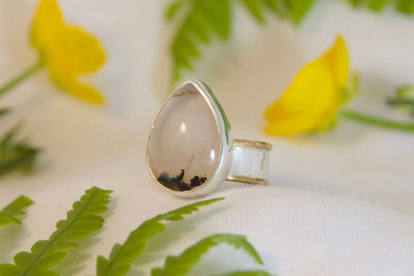 Silver ring with a teardrop-shaped stone on a white surface with green leaves and yellow flowers.