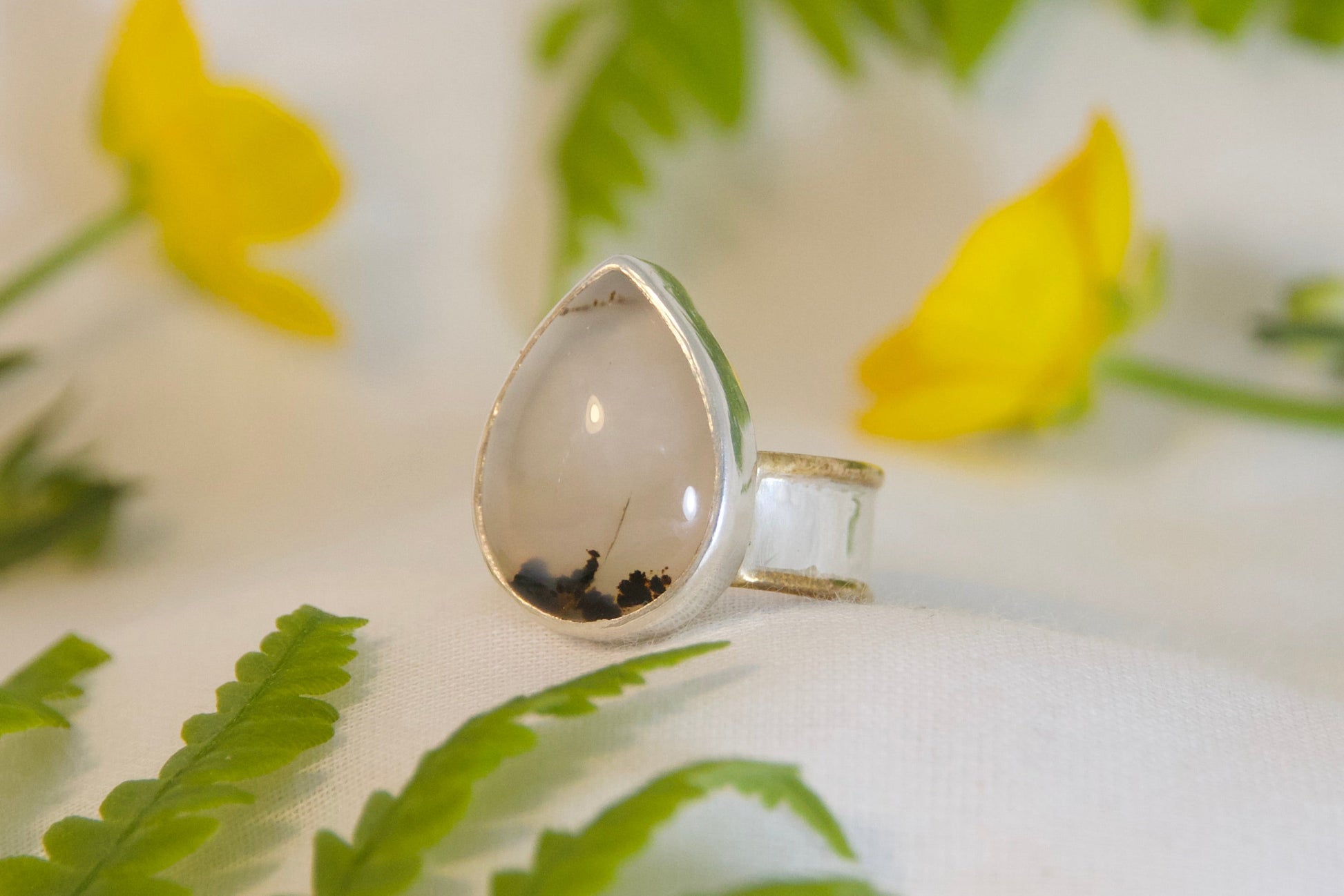 Silver ring with a teardrop-shaped stone on a white surface with green leaves and yellow flowers.