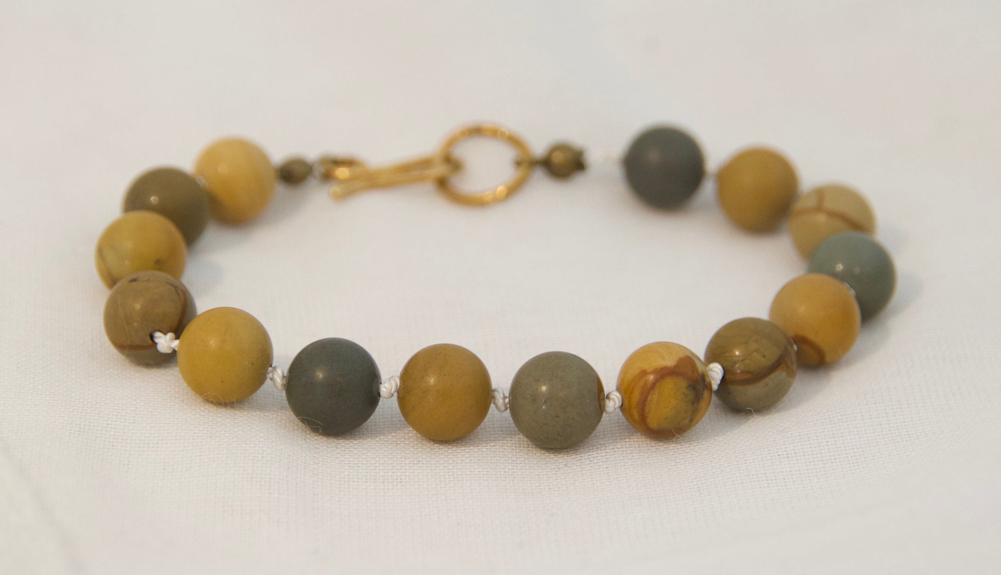 A beaded bracelet made of round stone beads in earthy brown, olive green, and gray tones, with a small gold clasp, displayed on a plain white background.