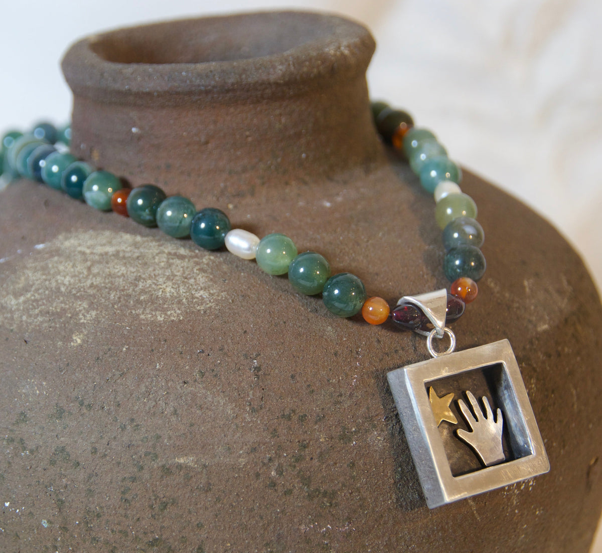 A beaded necklace with green, blue, and orange agate-like beads and a square silver pendant featuring a hand cutout, draped over a rustic brown ceramic vase against a soft white fabric background.