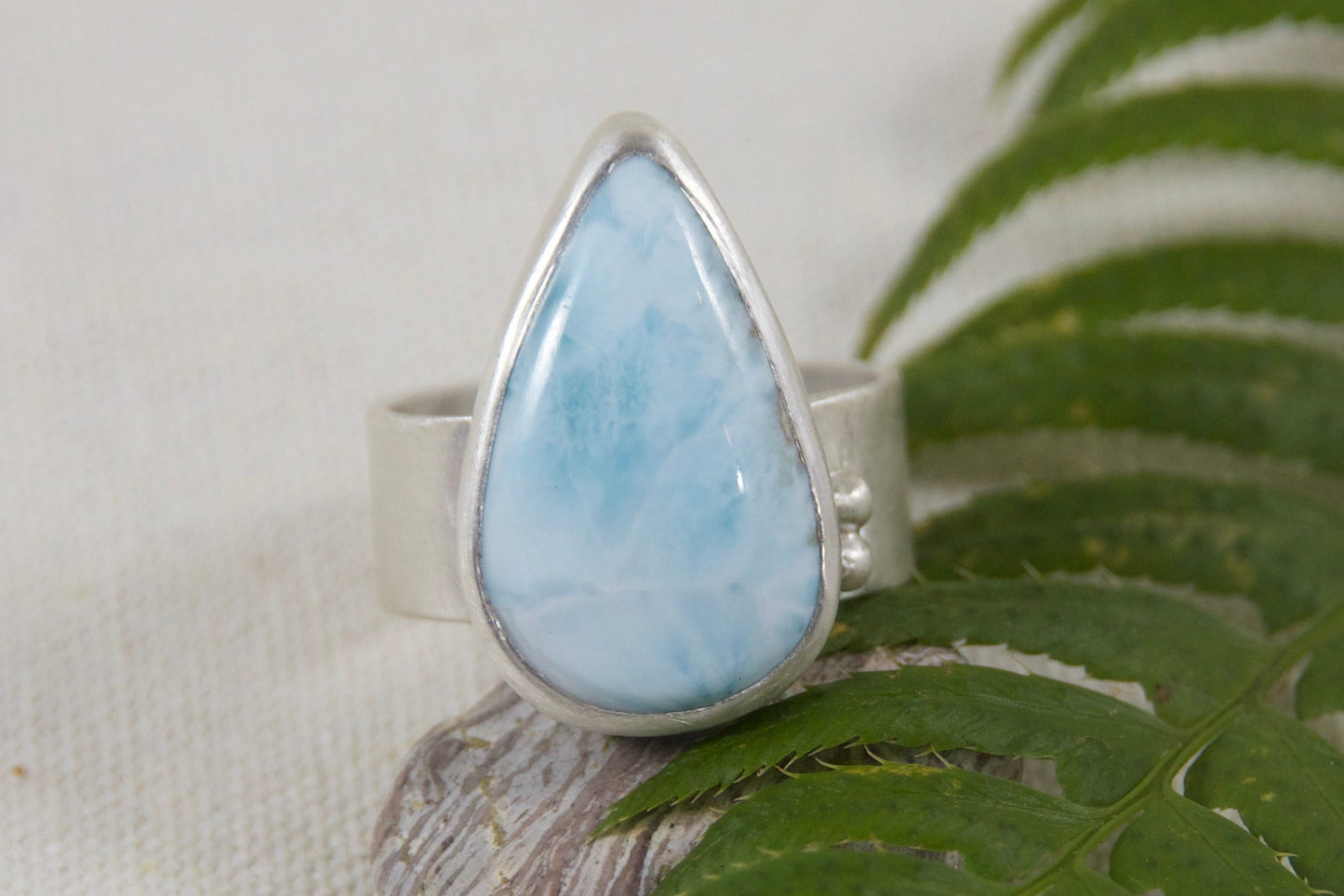 Teardrop-shaped gemstone ring on a green leaf against a beige background