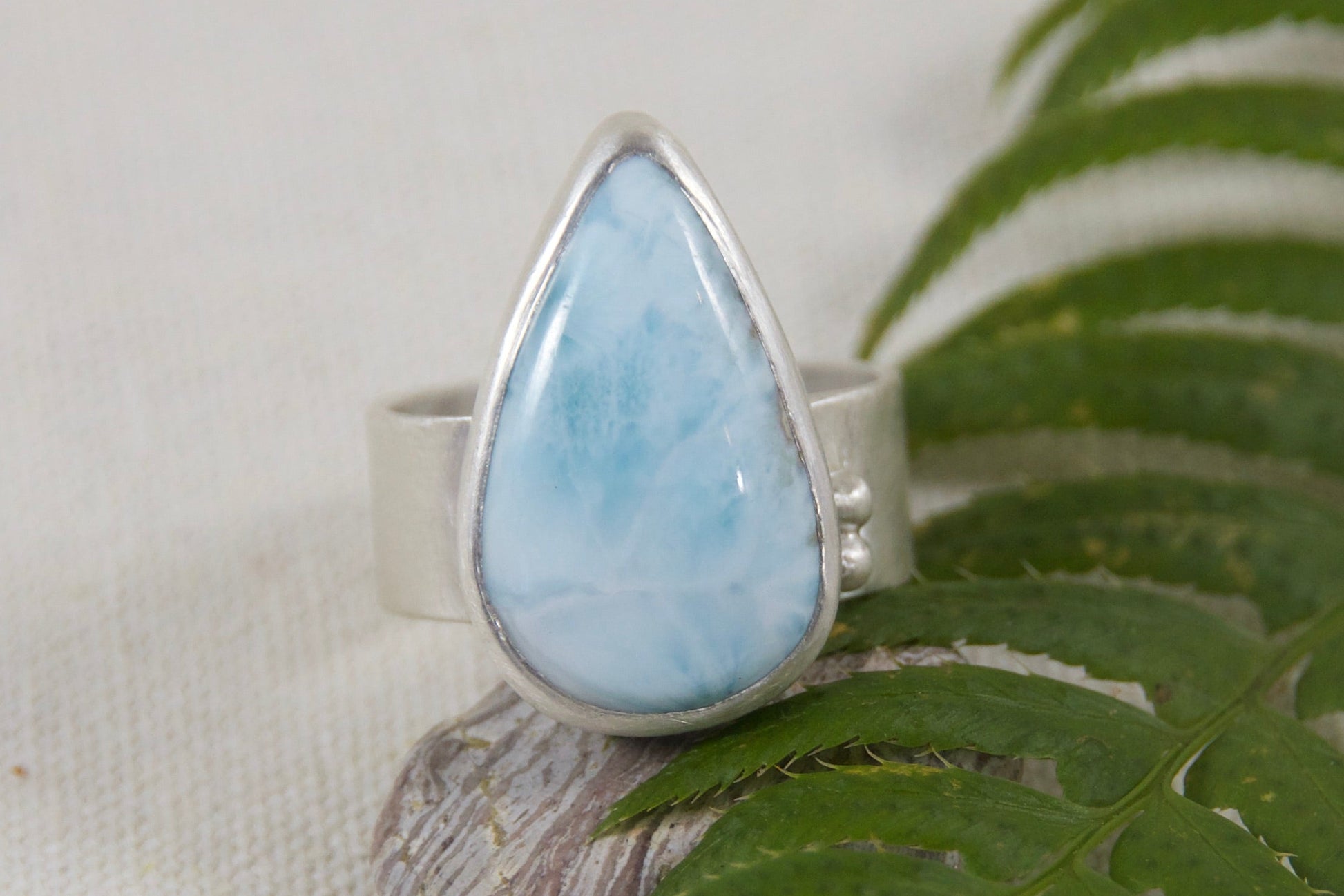 Teardrop-shaped gemstone ring on a green leaf against a beige background