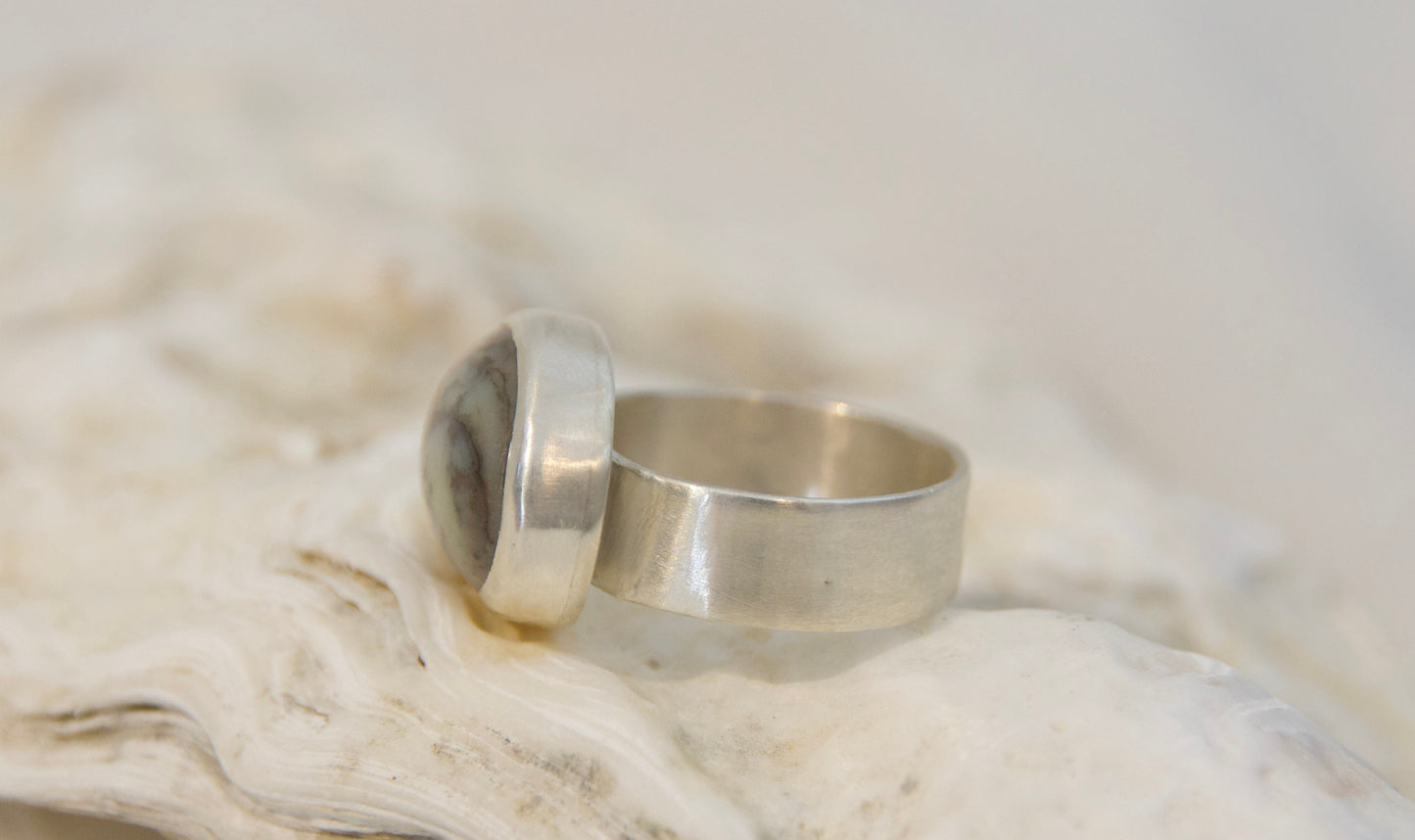 A silver ring with a large, polished oval stone, displayed on a textured white seashell against a soft, light background.