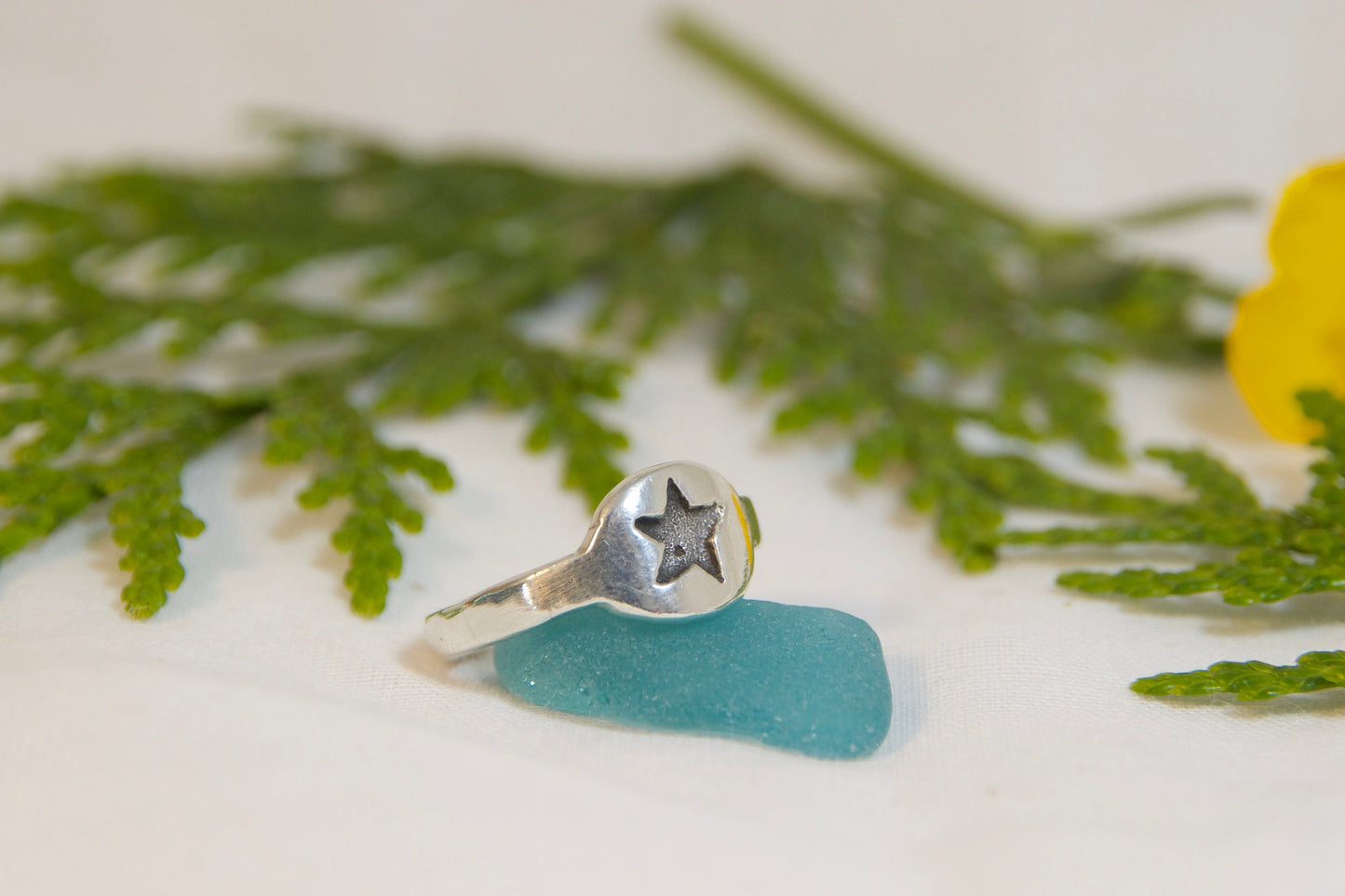 Silver ring with a star design on a piece of blue sea glass, surrounded by green leaves and a yellow flower.