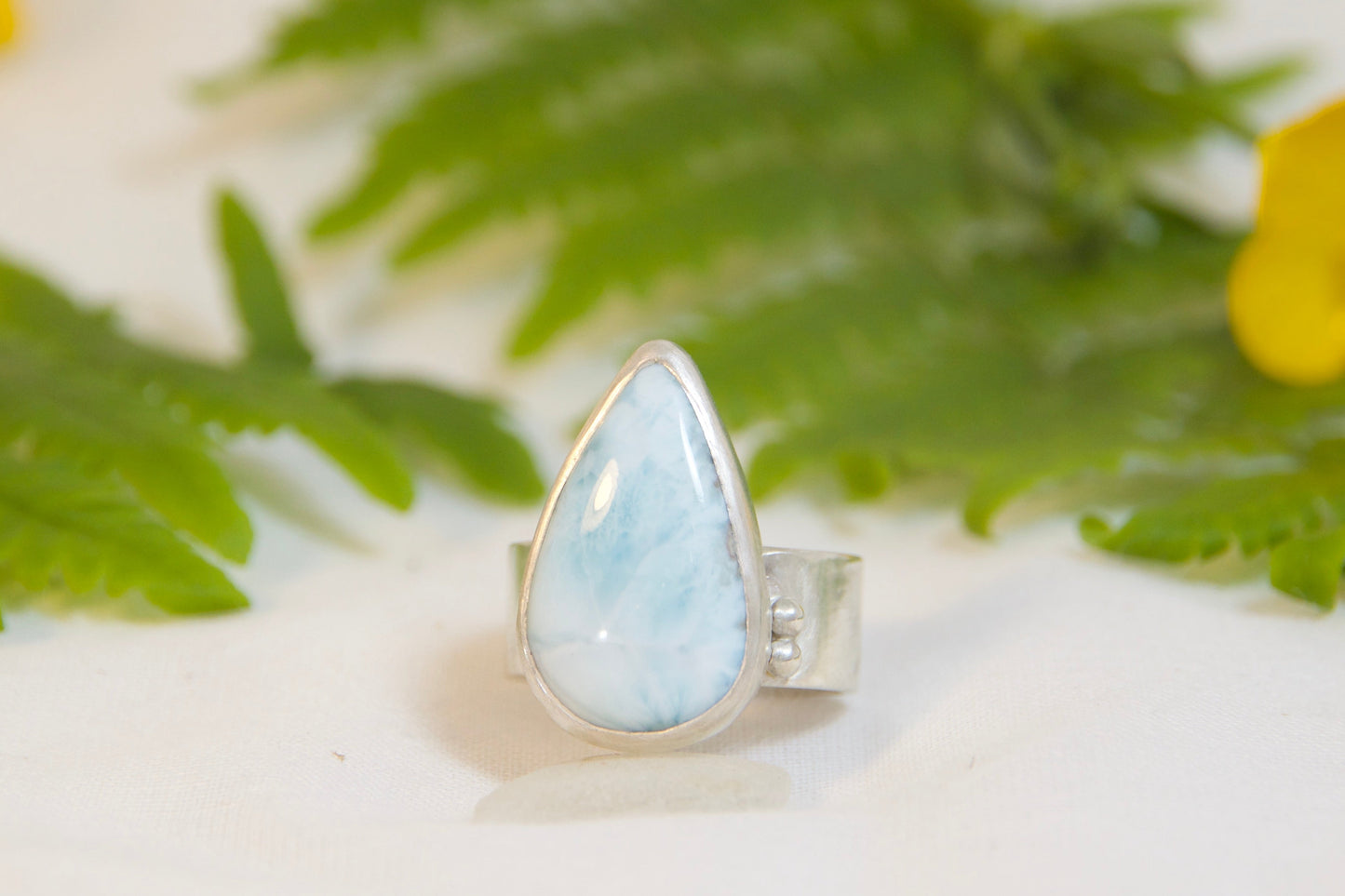 Ring with a teardrop-shaped gemstone on a white surface with green leaves and yellow flowers.