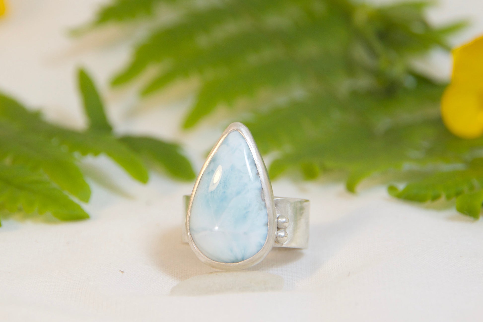 Ring with a teardrop-shaped gemstone on a white surface with green leaves and yellow flowers.