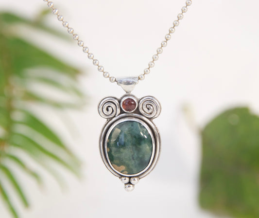 Silver necklace with a pendant featuring green stone and red gemstone against a blurred green leaf background