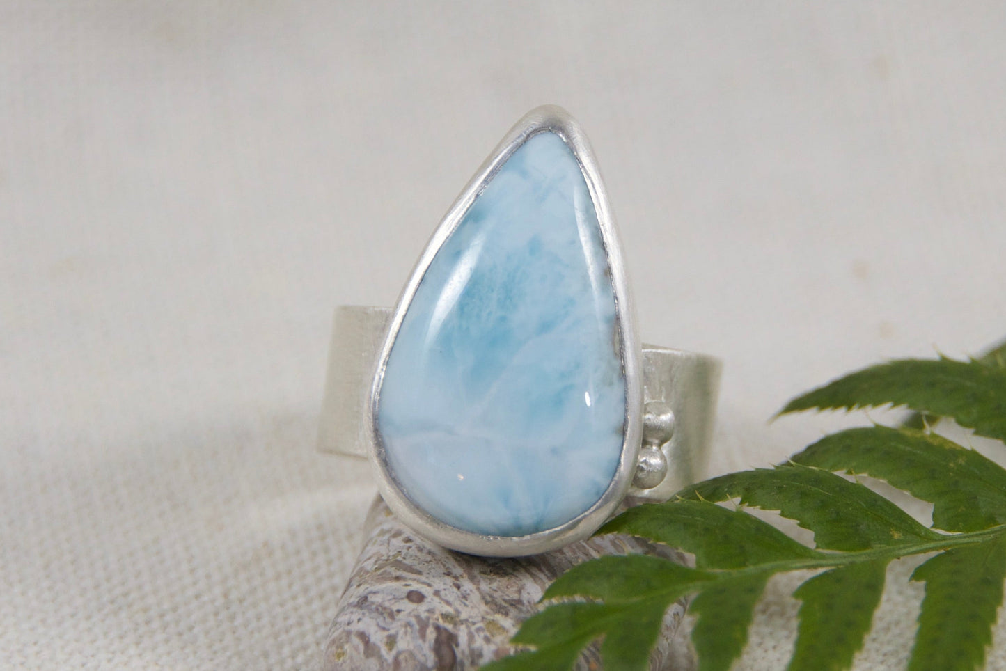 Teardrop-shaped larimar stone on a silver setting with a green leaf on a light gray background