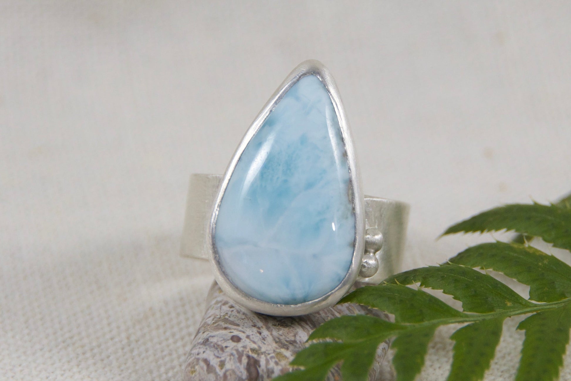 Teardrop-shaped larimar stone on a silver setting with a green leaf on a light gray background