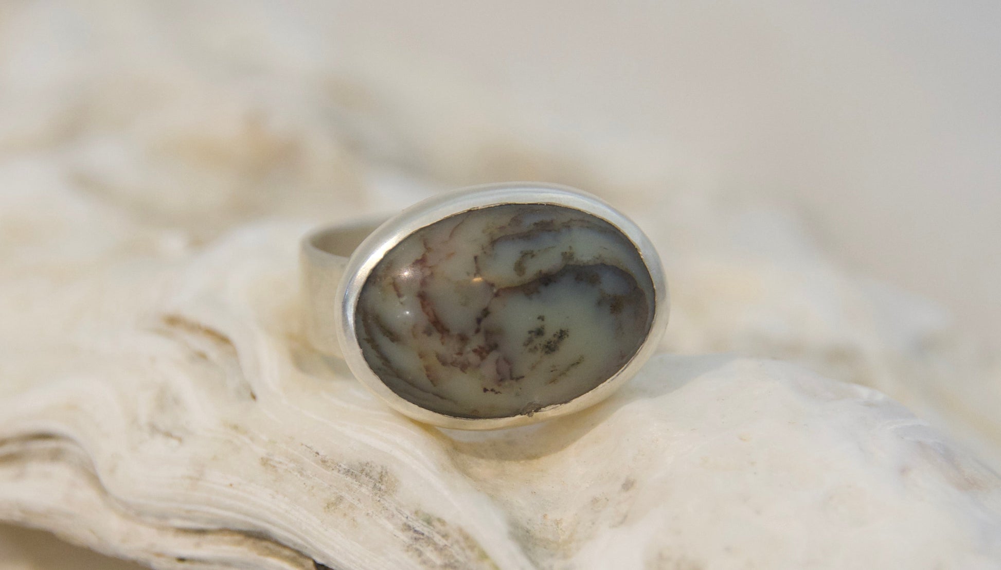 A silver ring with an oval cabochon stone featuring a mottled gray, brown, and white pattern, displayed on a textured white oyster shell.