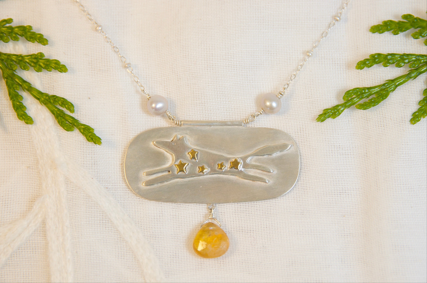 Necklace with a silver pendant featuring a dog design and a yellow gemstone, displayed on a white fabric background with green leaves.