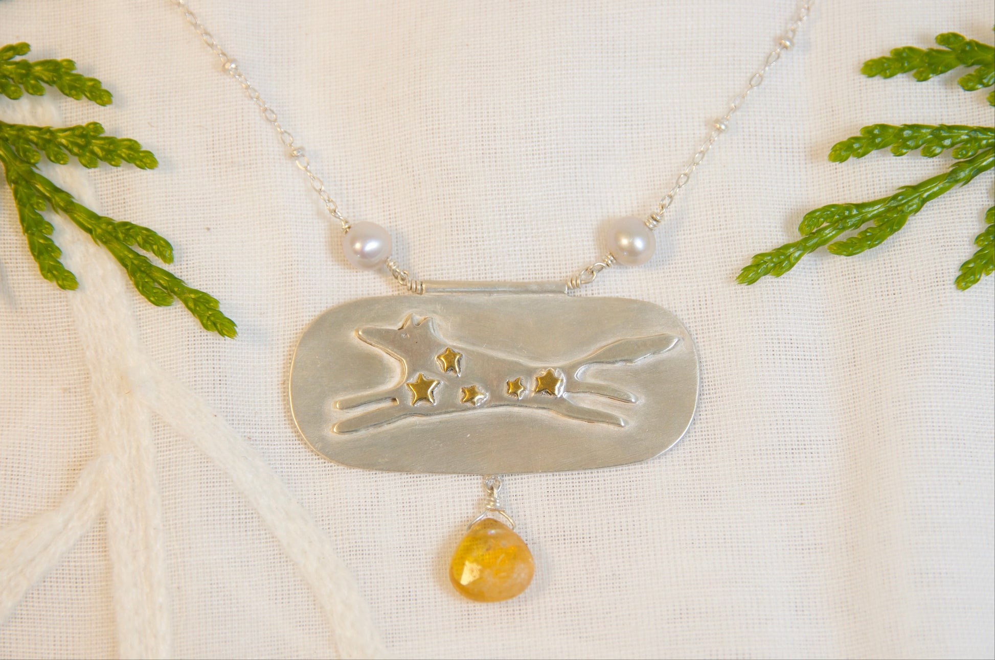 Necklace with a silver pendant featuring a dog design and a yellow gemstone, displayed on a white fabric background with green leaves.