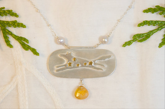 Necklace with a silver pendant featuring a dog design and a yellow gemstone, displayed on a white fabric background with green leaves.
