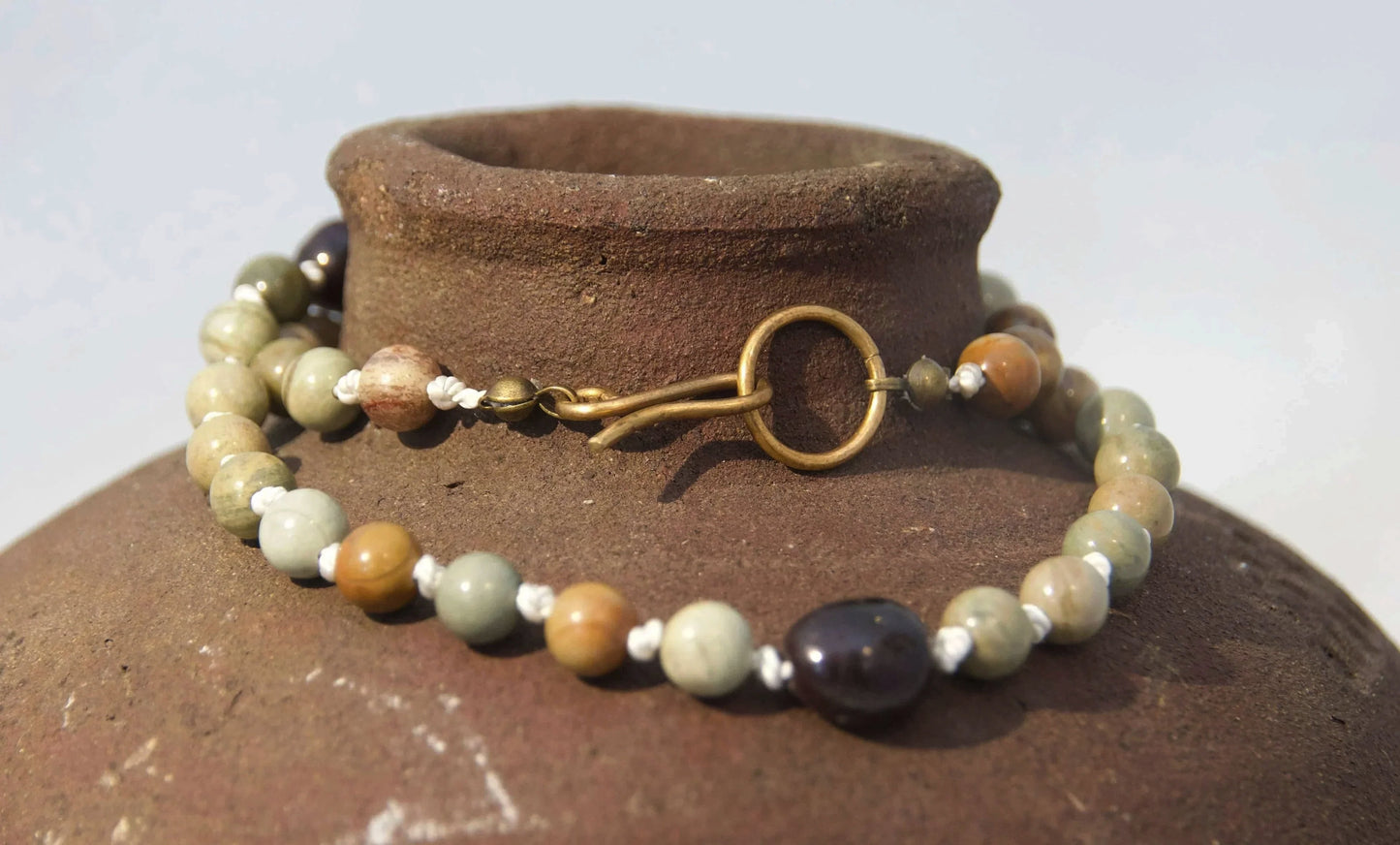 A beaded necklace with earthy green, brown, and white beads, featuring a gold-tone split ring clasp, draped around the neck of a rustic brown clay jar against a light gray background.