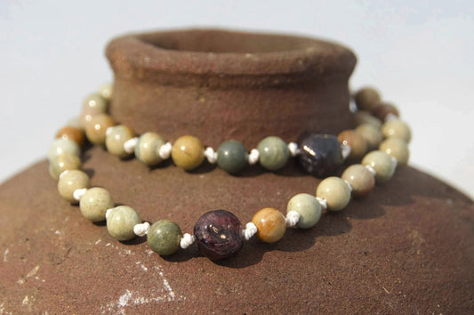 Hand knotted necklace with light green, yellow, and gray stone beads, displayed on a rustic brown ceramic vase against a white background. 