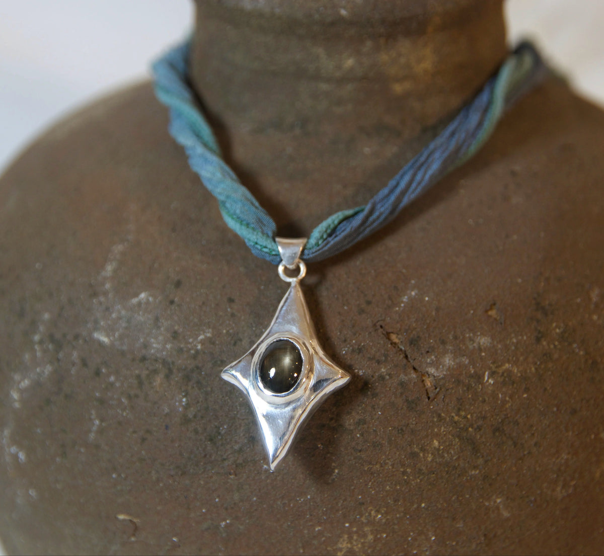 A silver pendant necklace with a four-pointed star design and a dark oval stone, displayed on a blue cord over a rustic brown ceramic vase.