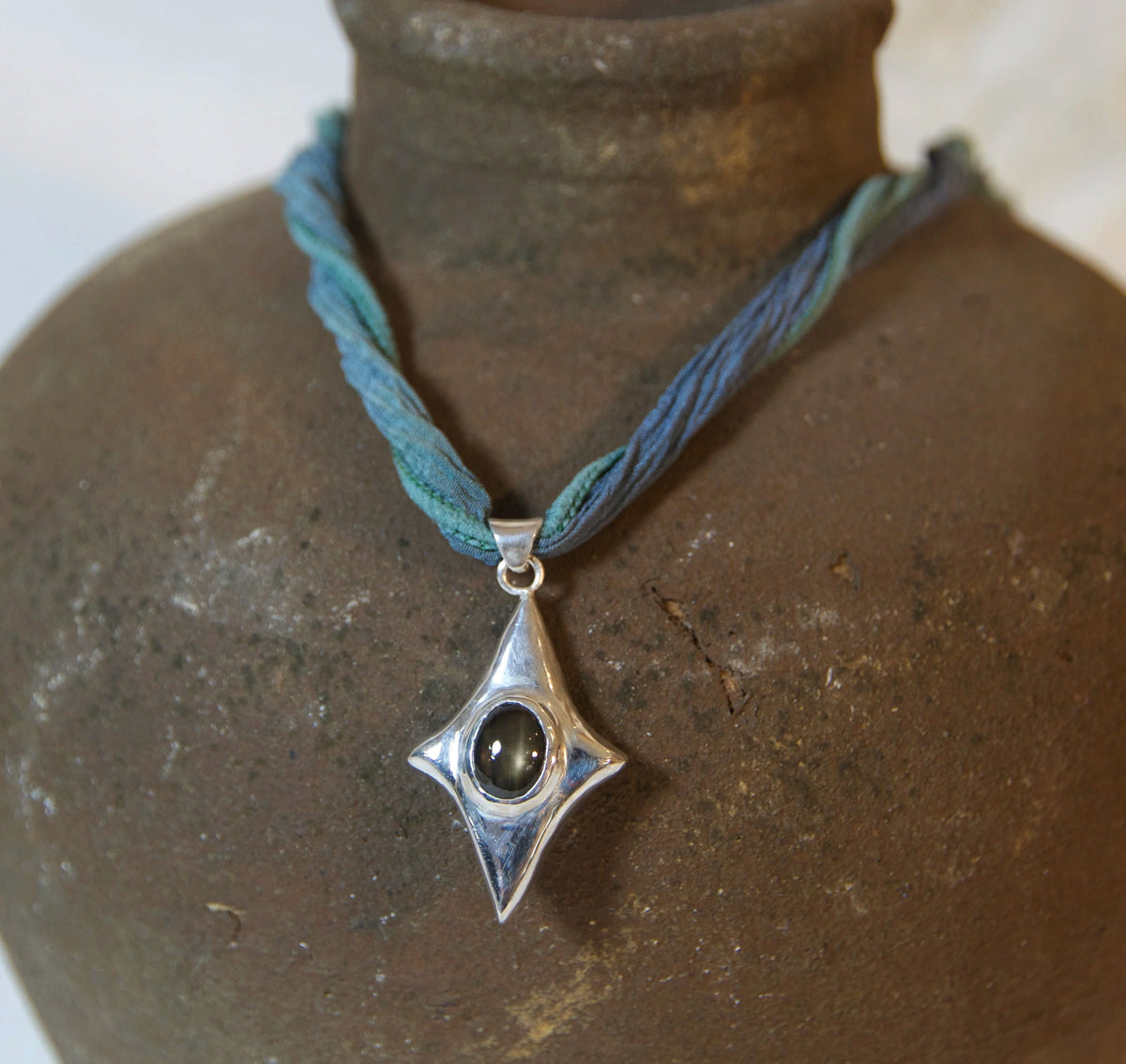 A silver pendant necklace with a blue and teal braided cord, displayed on a rustic brown ceramic vase against a soft, light beige background.