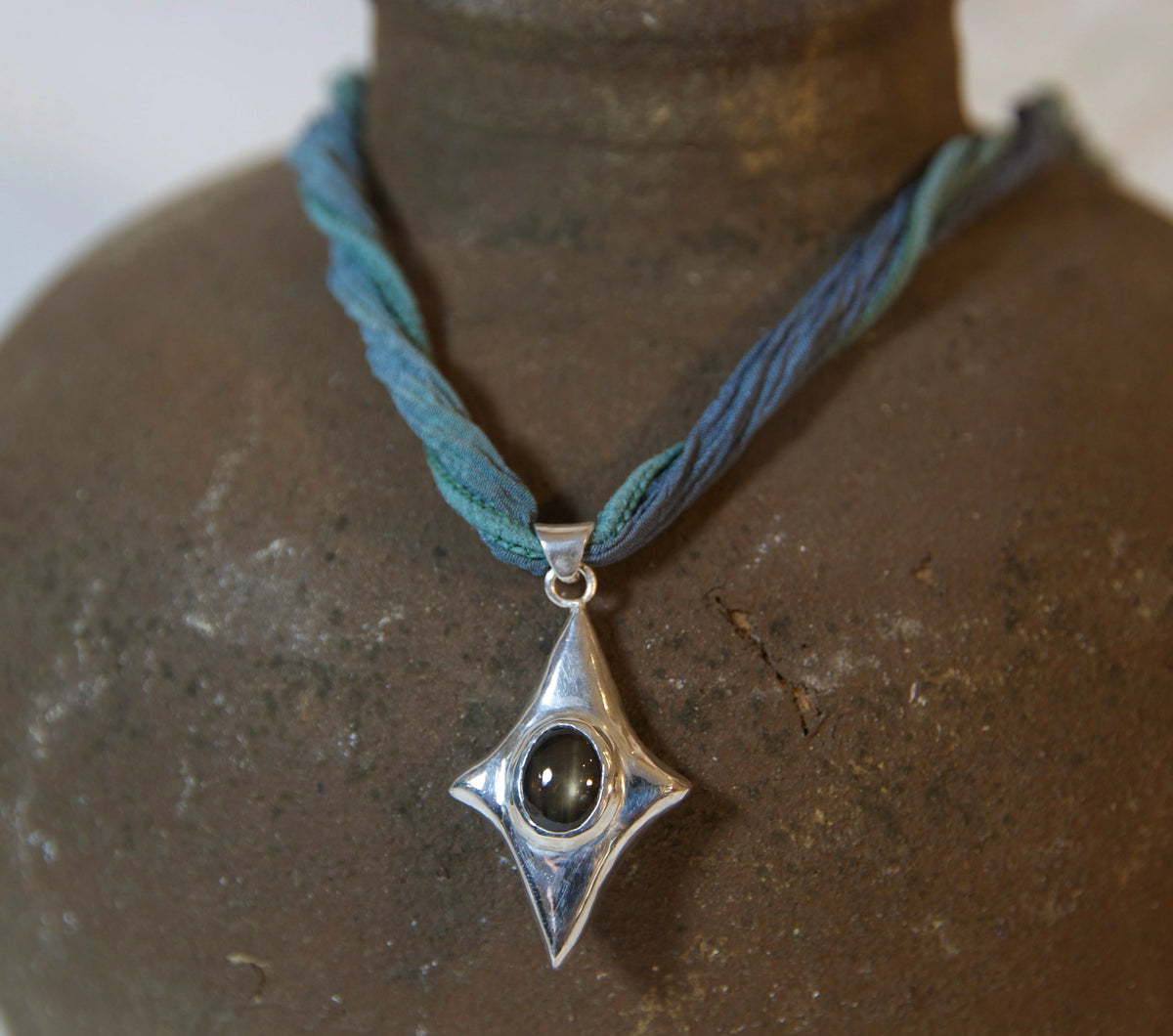 A silver pendant necklace with a blue braided cord, displayed on a rustic brown ceramic bust.