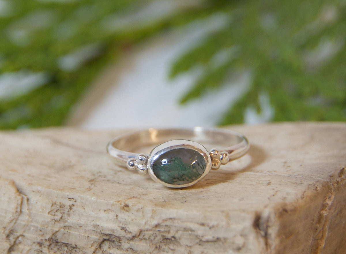 A silver ring with an oval green stone resting on a textured stone block, styled with green pine needles on a white fabric background.