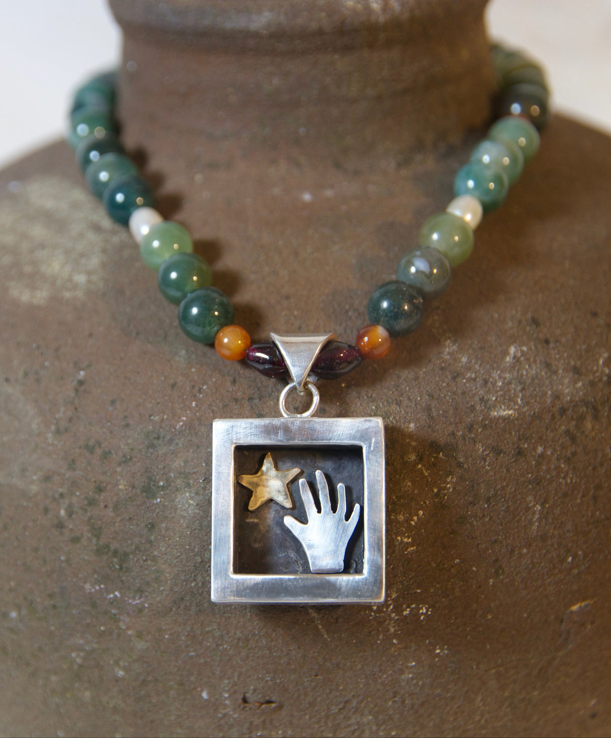 A beaded necklace with green, gray, and orange agate-like beads and a square silver pendant featuring a silver hand and a gold star, displayed on a rustic brown ceramic vase.