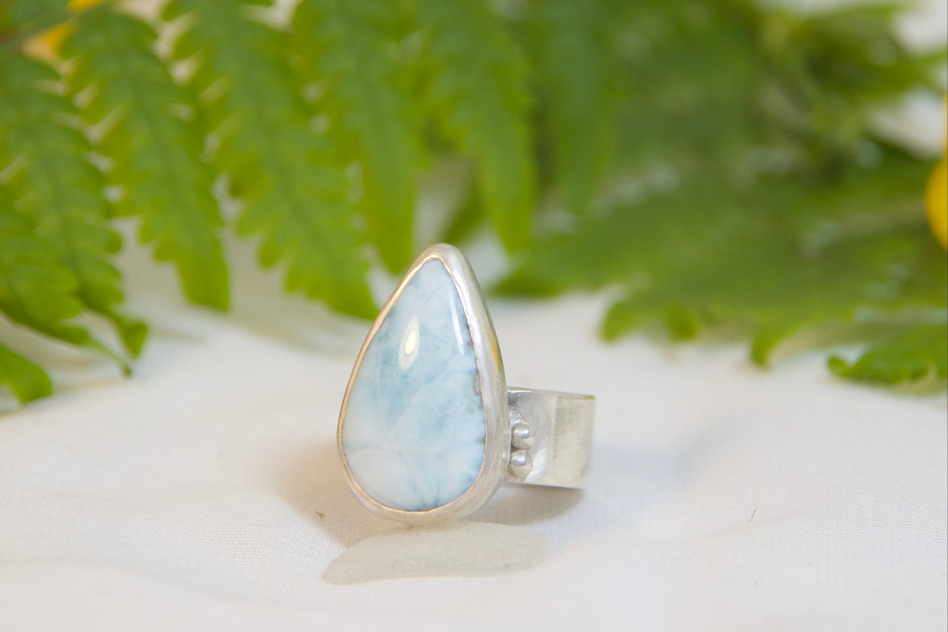 Silver ring with a blue stone on a white surface with green leaves and a yellow flower.