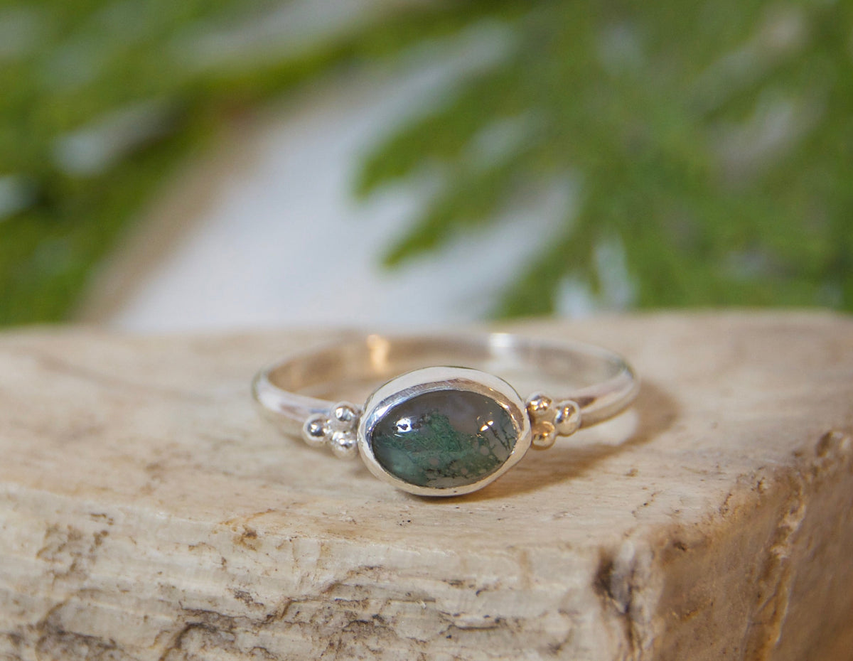 A silver ring with an oval green stone resting on a textured light beige stone, with green foliage in the background on a white fabric surface.