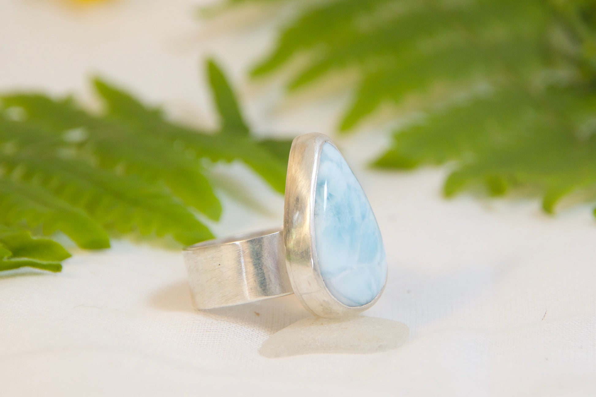 Silver ring with a blue stone on a white surface with green leaves and yellow flowers.