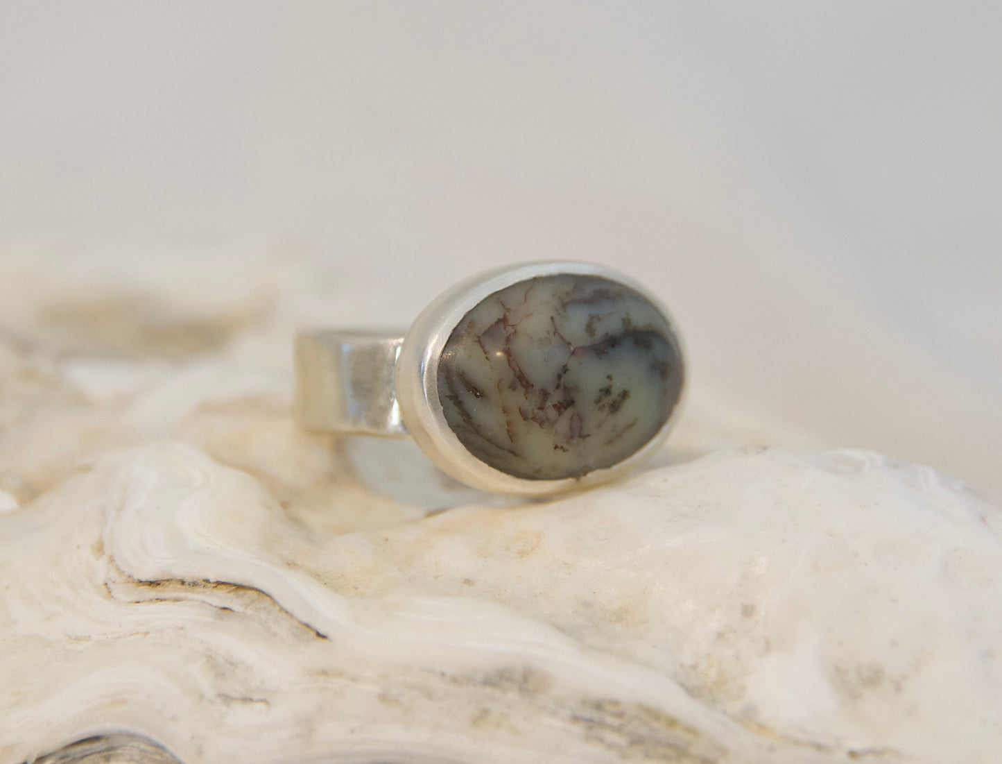 A silver ring with an oval cabochon gemstone featuring a gray and black marbled pattern, displayed on a textured white shell against a soft, light gray background.