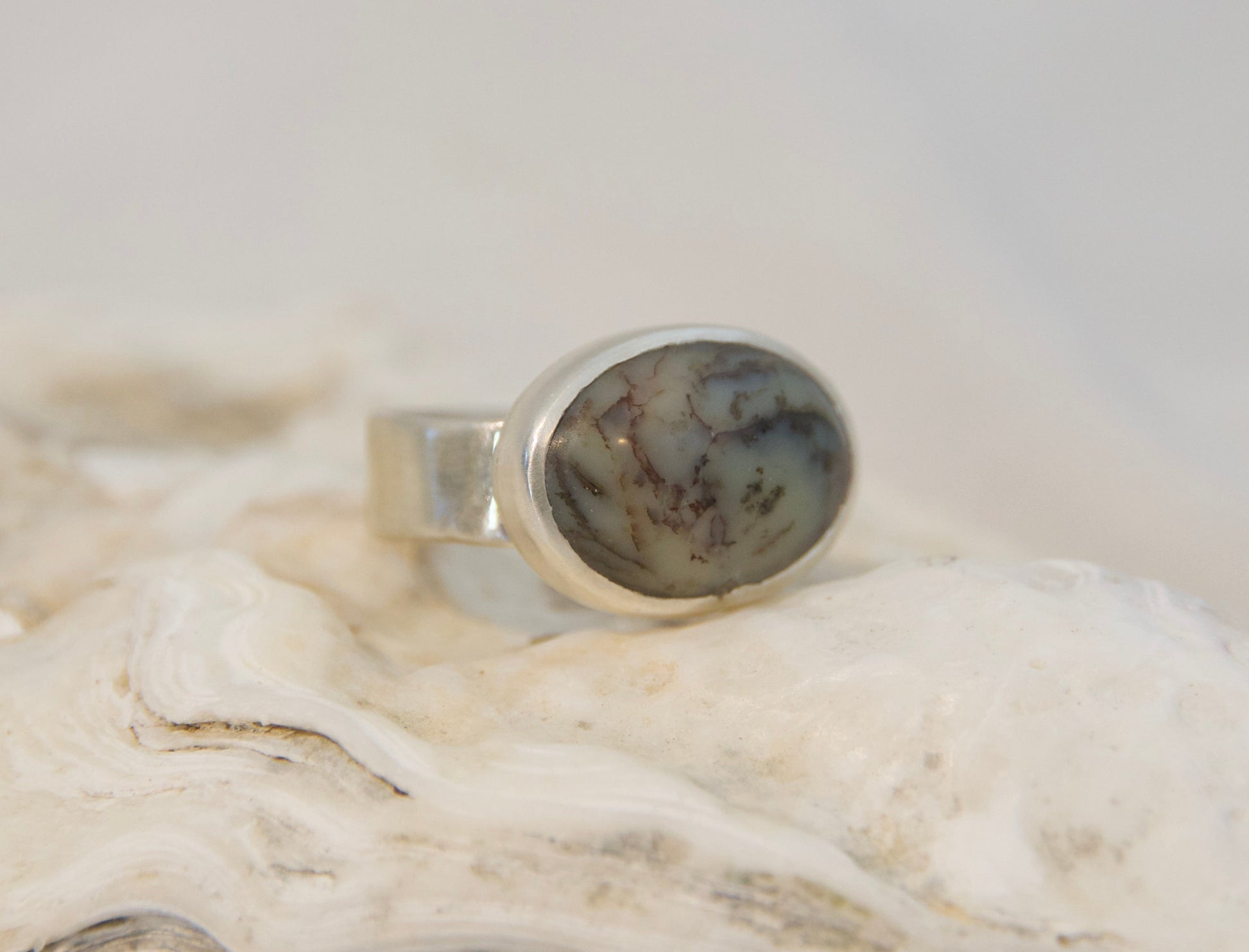 A silver ring with an oval cabochon gemstone featuring a gray and black marbled pattern, displayed on a textured white shell against a soft, light gray background.