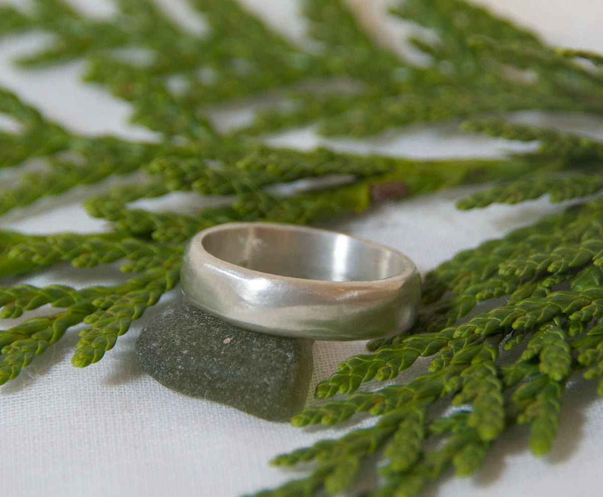 A simple silver band ring resting on a dark stone, placed on a green cedar branch over a white fabric background.