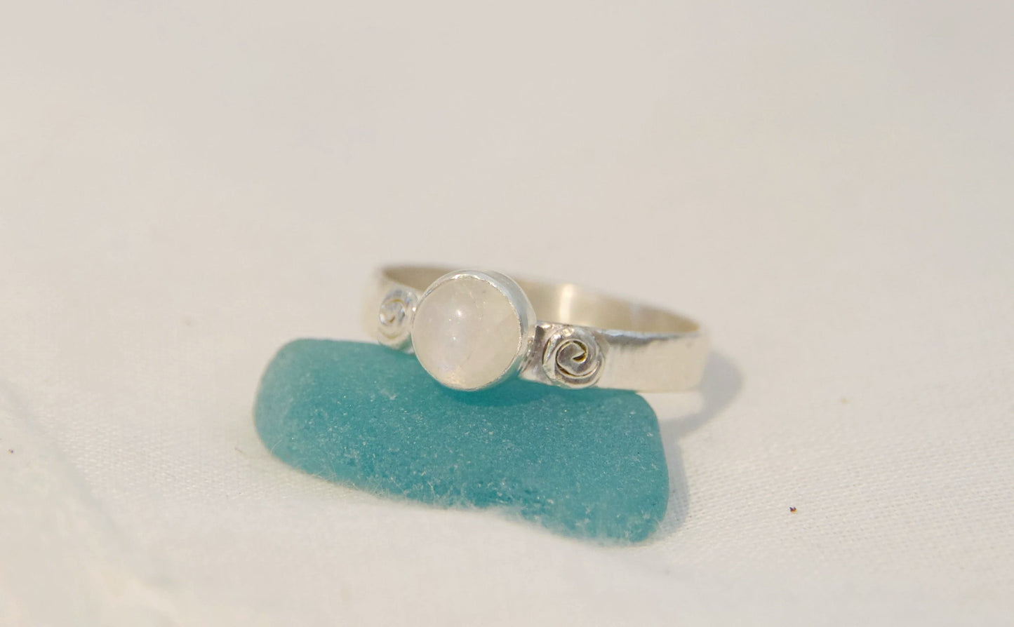 A silver ring featuring a round white moonstone center stone and swirly accents, resting on a piece of turquoise sea glass against a light off-white background.