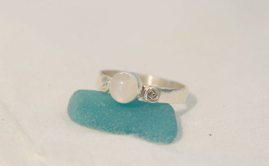 A silver ring featuring a round white moonstone center stone and swirly accents, resting on a piece of turquoise sea glass against a light off-white background.