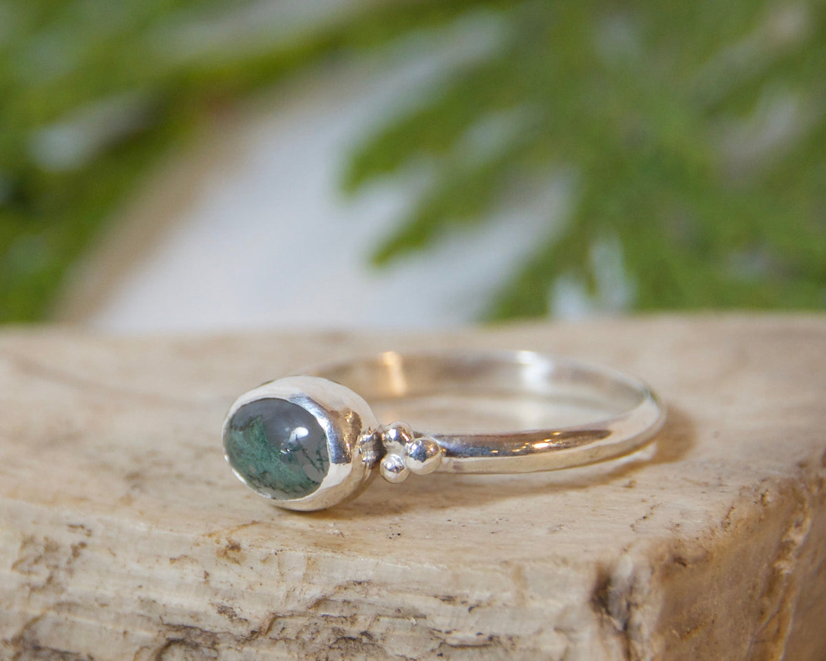A silver ring with a round green gemstone resting on a textured beige stone block, with green pine needles in the background on a white surface.