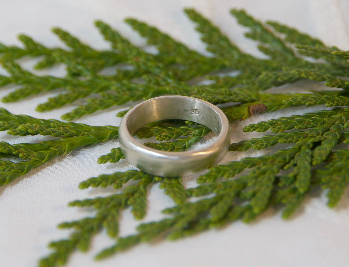 A simple silver band ring resting on a green branch over a white textured fabric background.