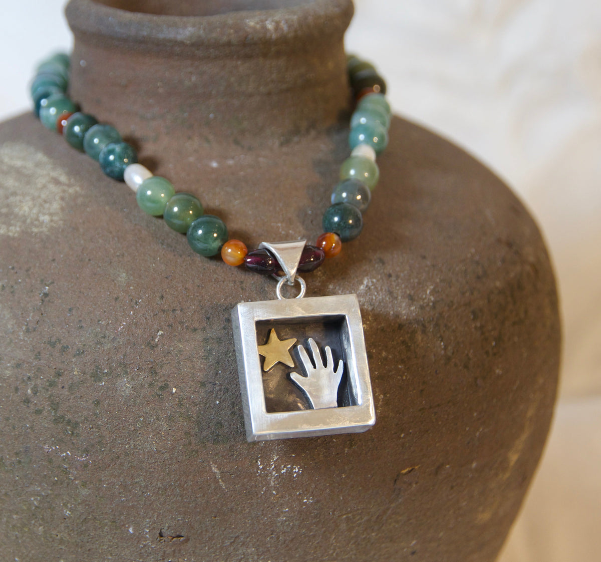 A beaded necklace with green, blue, and amber beads and a square silver pendant featuring a hand and star, displayed on a rustic brown ceramic vase against a soft white fabric background.