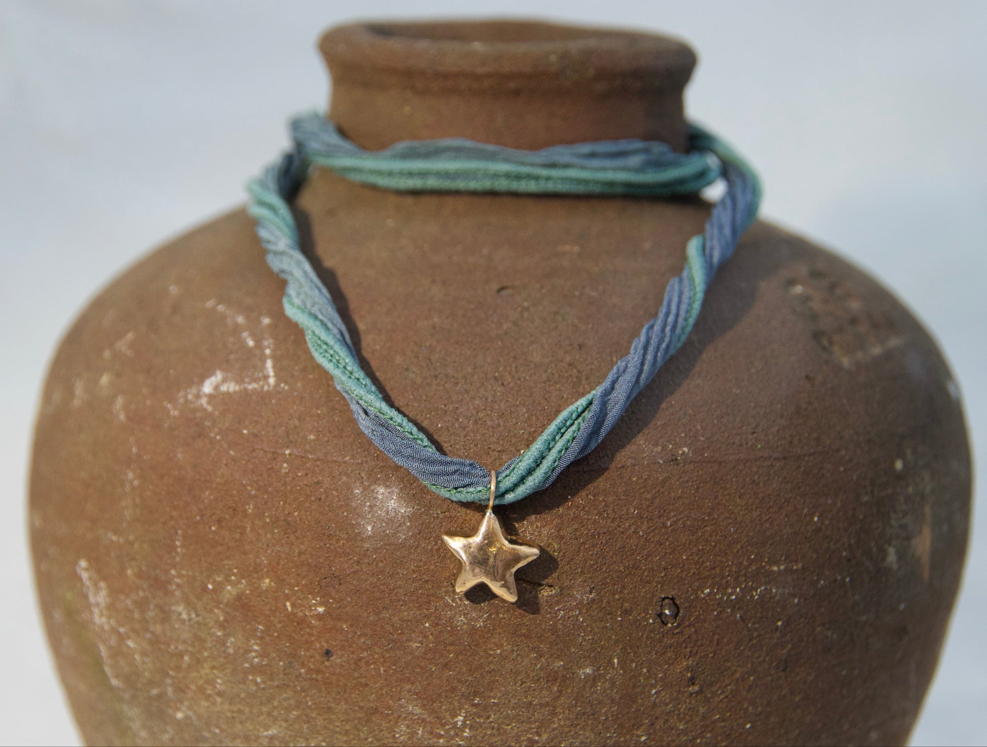 A blue and green braided cord necklace with a small bronze star pendant, displayed on a rustic brown clay vase against a light gray background.