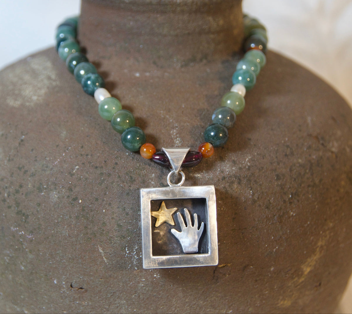 A beaded necklace displayed on a rustic brown ceramic vase against a soft white fabric background. The necklace features a mix of green, blue, and orange beads with a square silver pendant containing a white hand graphic and a gold star.
