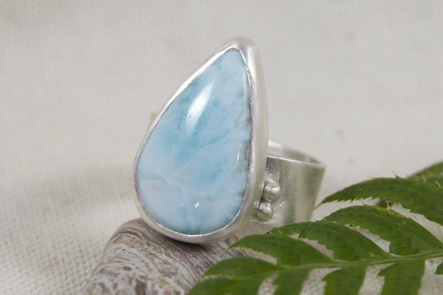 Silver ring with a blue teardrop-shaped gemstone on a white background with a fern leaf.