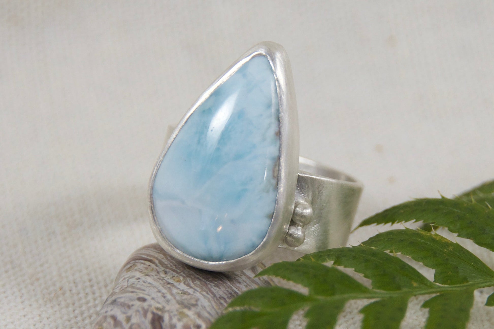Silver ring with a blue teardrop-shaped gemstone on a white background with a fern leaf.