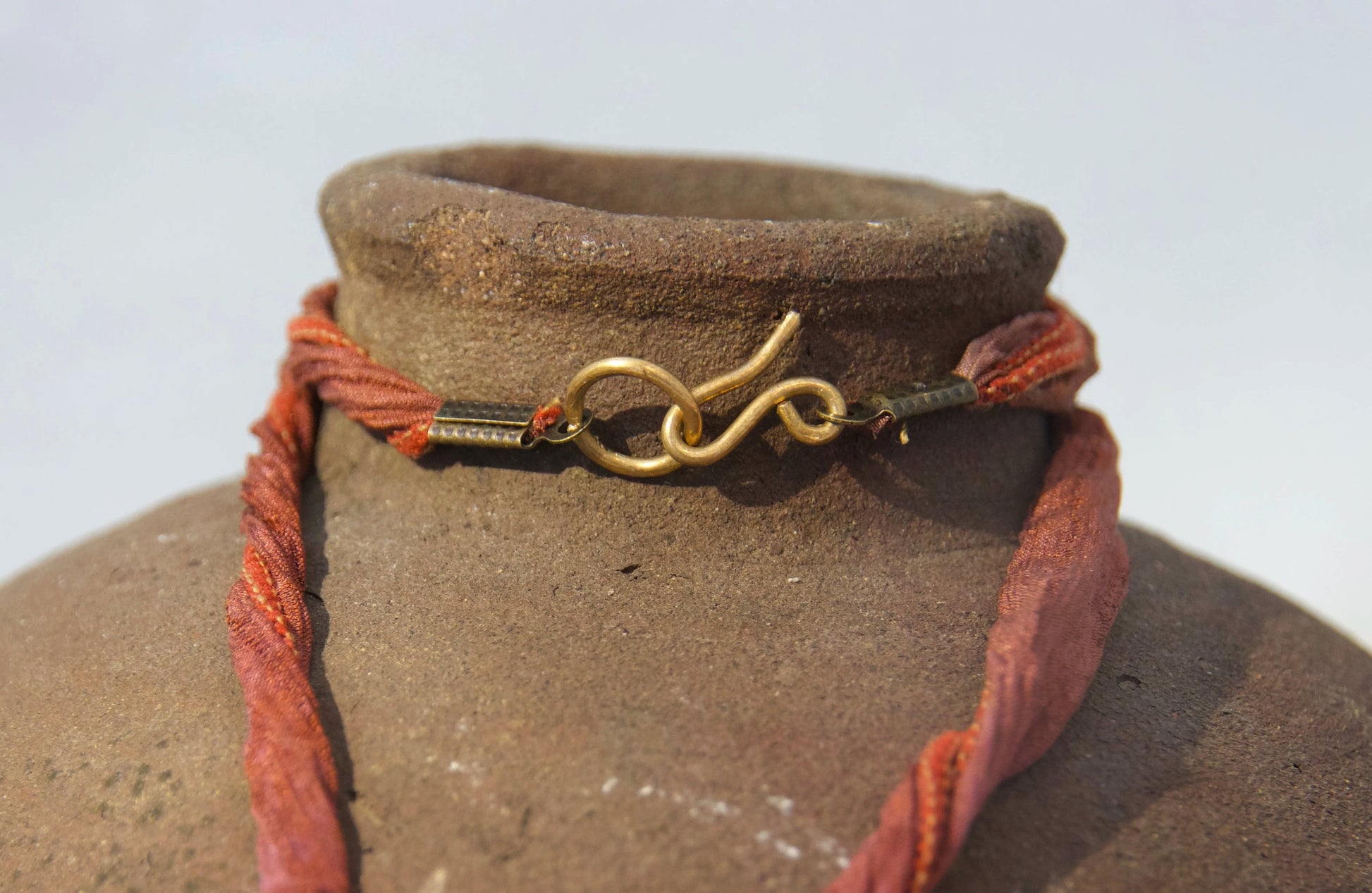 A close-up of a rustic brown ceramic vase or planter with a braided terracotta rope cord wrapped around its neck and a small brass handmade clasp, set against a bright white background.